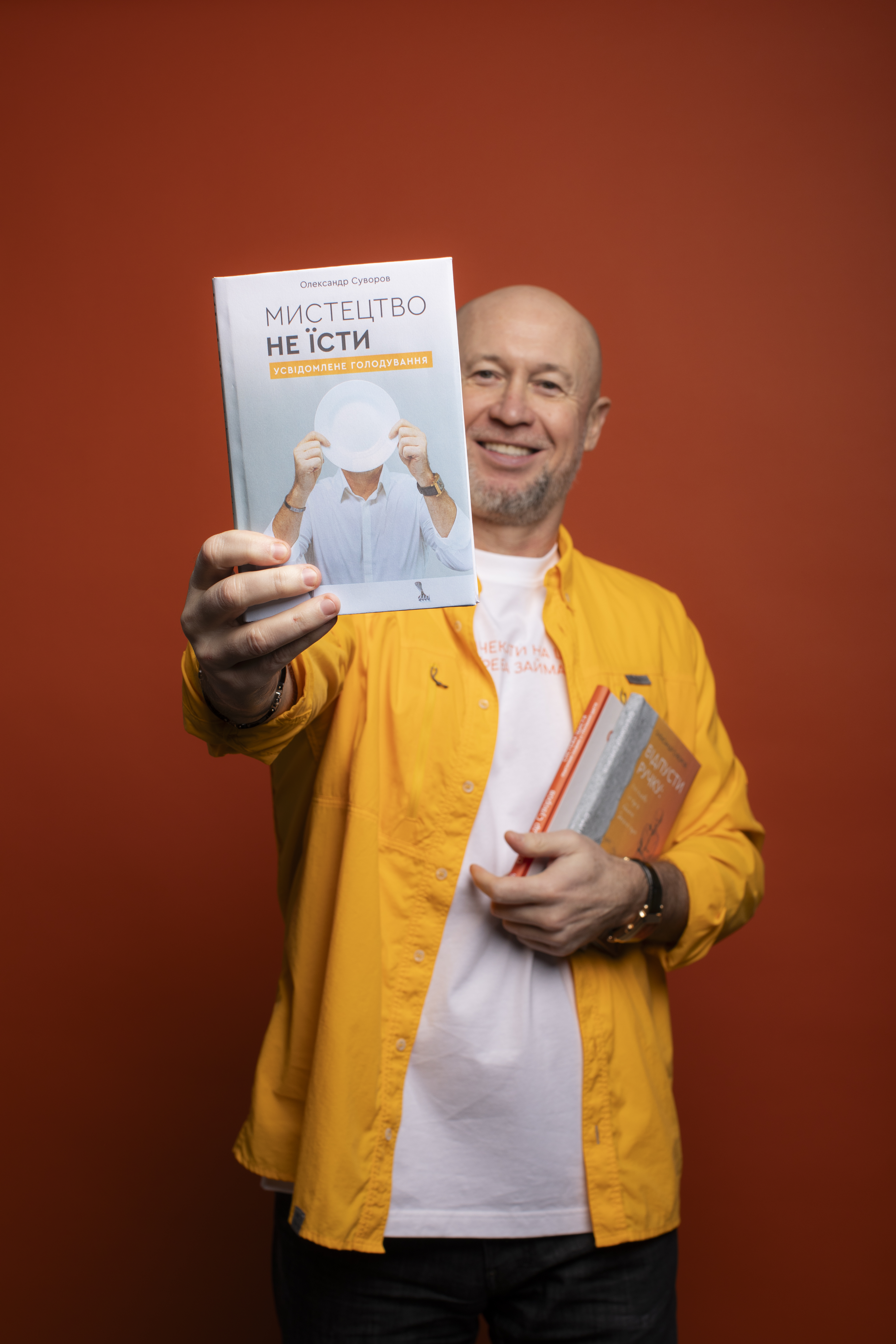 Oleksandr Suvorov is holding a book titled "The Art of Not Eating," which he authored. He is smiling and wearing a bright yellow shirt over a white T-shirt. In his other hand, he holds several more of his own books. The background is a solid orange color.