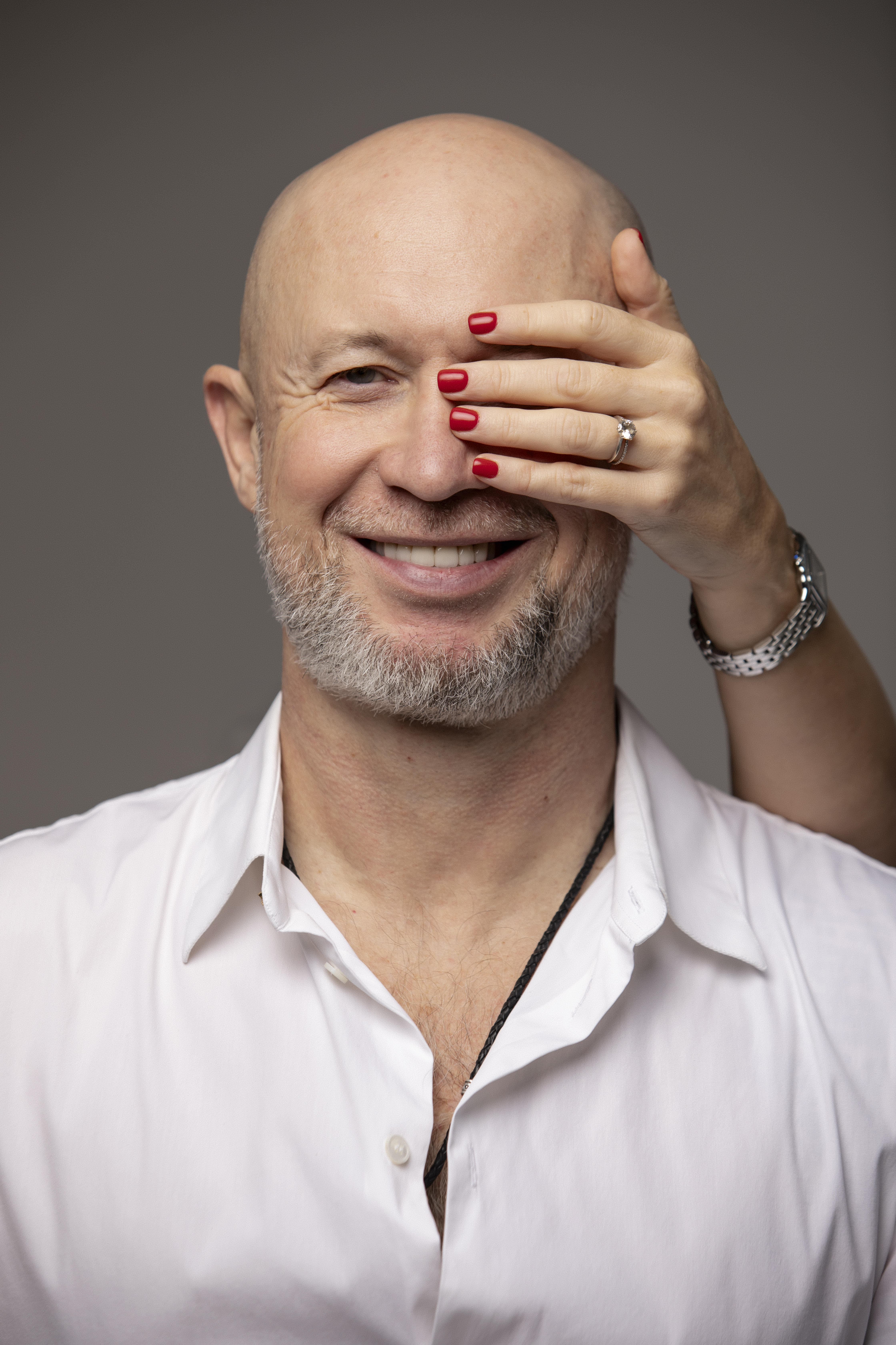 Oleksandr Suvorov, a man with a gray beard wearing a white shirt, is smiling. A woman's hand with red nail polish and a ring on the ring finger covers one of his eyes.